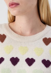 Pull en tricot avec un fond crème, arborant des cœurs en marron, jaune, vert et violet. Encolure côtelée, texture douce.