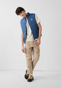 Chaleco acolchado azul sobre una camiseta crema, combinado con pantalones cargo beige y zapatillas marrones. Presenta un logo en el pecho del chaleco.