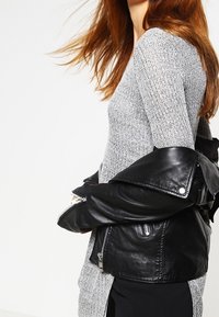 Giacca di pelle nera appoggiata su un maglione grigio a coste, caratterizzato da una vestibilità aderente e maniche lunghe. La giacca presenta dettagli visibili con zip.