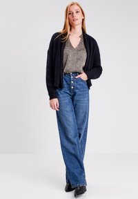 Cardigan nero sopra una maglietta a fantasia, abbinato a jeans blu a vita alta. I jeans presentano bottoni sulla parte anteriore. Texture complessiva liscia.