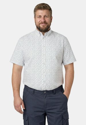 Smilende mand med kort skæg iført en hvid kortærmet skjorte med lille print og mørke cargoshorts, stående med den ene hånd i lommen.