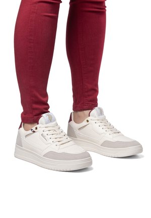 Sneakers bianchi e beige con una finitura testurizzata, caratterizzati da un accento sul tallone color bordeaux e un logo in rilievo, abbinati a jeans skinny rossi.
