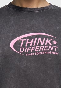 Κοντινή λήψη ενός σκούρου γκρι πουκάμισου με ροζ κείμενο «THINK DIFFERENT START SOMETHING NEW» και μια καμπυλωτή γραμμή με σχέδιο αστεριού πάνω από το κείμενο.