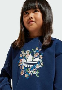 Svetlo modra majica s kapuco z cvetličnim vzorcem okoli belega logotipa Adidas. Ima mehak material in okroglo izrez. 