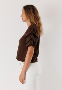 Top corto in cotone marrone con maniche a sbuffo, caratterizzato da una texture liscia. Indossato con jeans bianchi attillati, mostrando un profilo laterale.