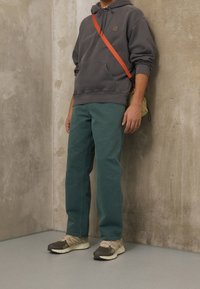 Persona che indossa una felpa grigia scura, pantaloni turchesi, sneakers beige e marroni e una borsa a tracolla beige con una tracolla arancione, in piedi contro un muro di cemento.
