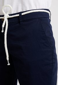 Pantalon bleu marine avec poche avant visible et passants pour ceinture, maintenu par une ceinture tressée blanche nouée en boucle.