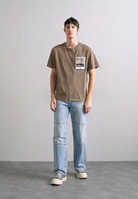 Pruun puuvillane T-särk, millel on graafiline tasku, koos helesiniste denimihallide teksadega, millel on lõdva lõikega ja nähtavad õmblused, ning valged tennisjalanõud.