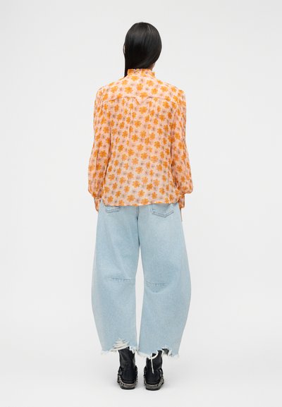 Γυναίκα με μακριά μαύρα μαλλιά, ντυμένη με πορτοκαλί μπλούζα με λουλούδια, ανοιχτό μπλε φαρδιά τζιν με σκισίματα και μαύρες μπότες, που στέκεται γυρισμένη με την πλάτη.