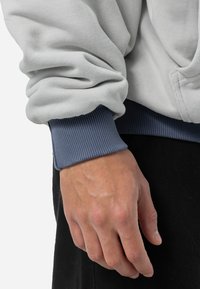 Sweatshirt gri deschis cu manșete ribbate de culoare bleumarin, evidențiind textura moale a materialului și o croială lejeră. Mână odihnindu-se aproape de detaliul cusăturii verticale.