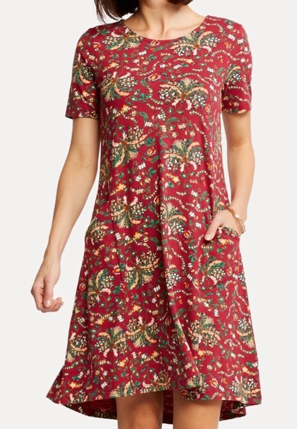 Vrouw in een korte mouw, knielange rode jurk met een bloemenpatroon en zakken, staand tegen een witte achtergrond.