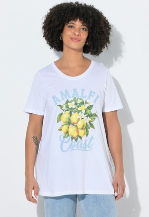 Frau mit lockigem Haar, die ein weißes T-Shirt mit der Aufschrift "Amalfi Coast" und einer Grafik eines Zitronenzweigs trägt, kombiniert mit hellblauen Jeans.