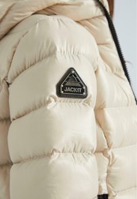 Helle beige Pufferjacke mit gestepptem Design, ausgestattet mit einem schwarzen dreieckigen Logopatch mit der Aufschrift "JACKIT" auf der oberen linken Seite.