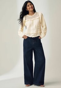 Kvinde står med lukkede øjne, iført en cremet langærmet bluse med blonde og mørke brede jeans med hænderne i lommerne.
