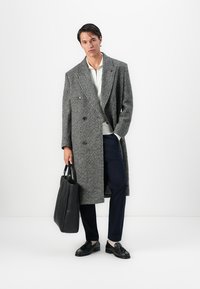 Mann in einem langen grauen Fischgrätenmantel, weißem Hemd, marinefarbener Hose, schwarzen Quastenlacken, mit einer großen schwarzen Leder-Tote-Bag in der Hand, vor einem weißen Hintergrund stehend.