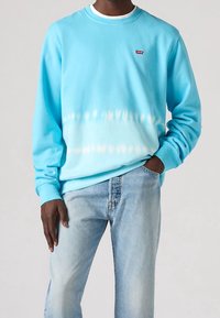 Sudadera azul claro con degradado tie-dye blanco, cuello redondo acanalado y pequeño logo rojo de Levi's en el pecho; combinada con vaqueros azul claro.