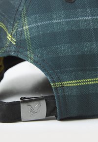 Casquette de baseball en tissu écossais vert foncé avec des accents jaunes. Elle présente un clip en métal argenté avec un motif d'aigle en relief.