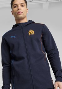Felpa con zip navy in tessuto morbido, con dettagli arancioni e blu, logo Puma e insegna di Marsiglia sul petto.