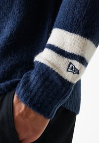 Jersey de punto azul marino con un acabado texturizado, que presenta una franja blanca y un parche con el logo en el puño. Primer plano del brazo y la manga.