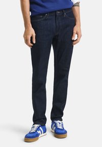 Dark blue denim jeans with a slim fit, featuring orange stitching and a classic five-pocket design, paired with blue athletic sneakers.