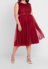 Rode mouwloze jurk met kanten lijfje en tule rok, met een getailleerde taille en doorzichtige overlay. Draag met nude hoge hakken.