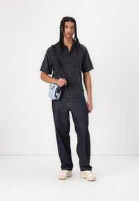 Kurzärmeliges schwarzes Shirt, dunkelblaue Jeans und cremefarbene Sneaker. Hält eine kleine hellblaue Umhängetasche mit schwarzen Akzenten.