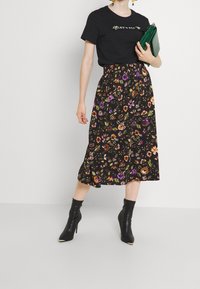 Black t-shirt features green text, paired with a floral midi skirt in black, orange, and purple. Accessorized with a green handbag and black ankle boots.