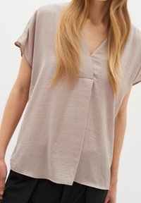 Ljus taupeblus i texturerad tyg, med V-ringning, korta ärmar och asymmetrisk fåll. Minimalistisk design med subtila detaljer.