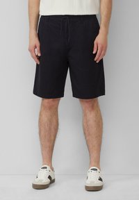 Pantalones cortos negros de tela ligera, con una cintura elástica con cordón, bolsillos laterales y un corte recto. Combinados con zapatillas blancas.