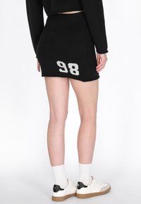 Donna che indossa una mini gonna nera a maglia con il numero "98" bianco sul lato, abbinata a calze bianche e sneakers bianche con dettagli neri.