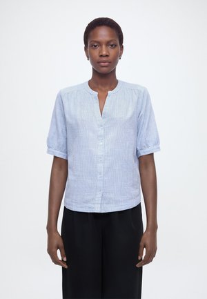 Vrouw met kort haar, gekleed in een lichtblauw overhemd met korte mouwen en zwarte broek, staand tegen een effen witte achtergrond.