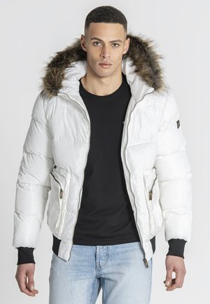 Mann trägt eine weiße Daunenjacke mit pelzgefütterter Kapuze, schwarzes Hemd und hellblaue Jeans, steht vor einem schlichten Hintergrund.