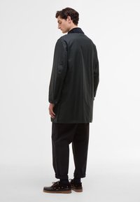 Casaco longo verde-escuro em tecido leve, com colarinho preto e punhos abotoados. Combinado com calças pretas e sapatos castanhos.