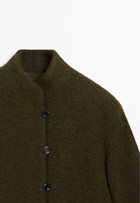 Přes oversized zelený vlněný kabát s texturovaným povrchem, který má stojáček a čtyři černé knoflíky vpředu.