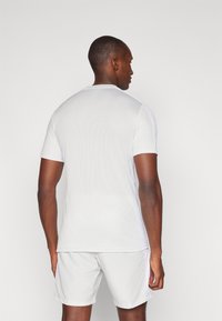 Maglietta sportiva bianca a maniche corte realizzata in materiale testurizzato e traspirante, con un design a girocollo, indossata con pantaloni corti abbinati.