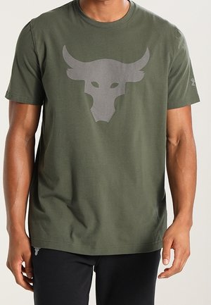 Mann trägt ein olivgrünes T-Shirt mit einem großen grauen Stierkopf-Logo auf der Brust und schwarze Hosen.