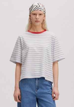 Femme portant un t-shirt blanc rayé avec un col rouge, un jean bleu, de grandes boucles d'oreilles et un foulard blanc à motifs sur fond uni.
