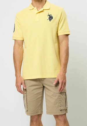 Polo jaune à manches courtes avec un tissu texturé. Il comporte un emblème bleu marine sur le côté gauche de la poitrine. Associé à un short cargo beige.