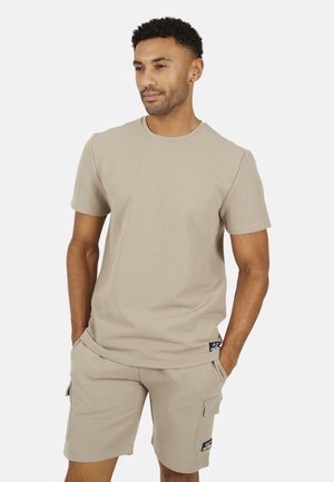 Uomo con capelli corti e ricci che indossa una camicia beige a maniche corte con texture e pantaloncini cargo abbinati, mani nelle tasche, che guarda a destra.