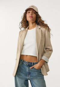 Béžový blazer s vyrolovanými rukávmi cez biely cropped top, skombinovaný so svetlo modrými džínsami s vysokým pásom. Klobúk je svetlo béžový s červeným textom.