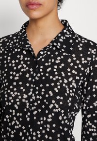 Chemise noire avec col, ornée d'un motif abstrait blanc. Fabriquée en tissu léger, avec des boutons sur le devant et une coupe décontractée.