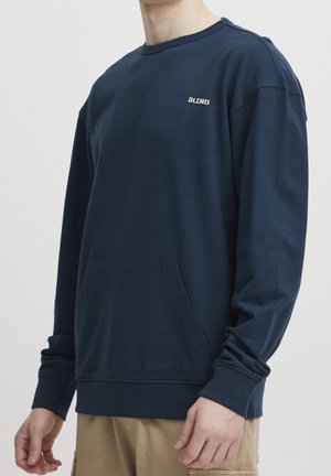 Homme portant un sweat-shirt bleu marine à manches longues avec poche avant et logo « BLEND » sur la poitrine, associé à un pantalon beige.