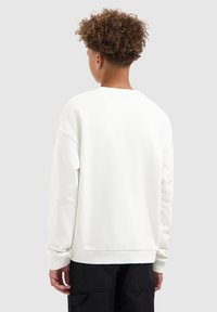 Jongere met krullend haar, gekleed in een eenvoudige witte sweatshirt en zwarte broek, gezien van achteren tegen een grijze achtergrond.