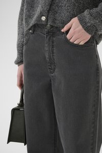 Hoogwaardige grijze spijkerbroek met een relaxed fit, voorzien van zijzakken en een gepolijste zilveren knoop. Een kleine groene handtas wordt vastgehouden.