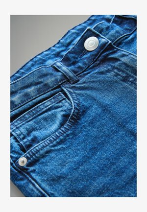 Siniset farkut, joissa on klassinen muotoilu, etutasku ja valkoinen nappi vyötäröllä, näyttäen hieman haalistuneelta tekstiurilta.