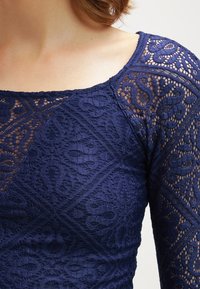 Donna che indossa un top in pizzo blu navy con motivi floreali e geometrici, caratterizzato da una manica in pizzo trasparente e un ampio scollo.