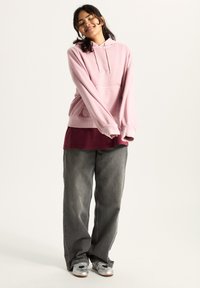 Pink hoodie with a front pocket, paired with a maroon layered top, grey baggy jeans, and silver shoes, standing against a light background.
