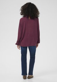 Burgundy long-sleeve blouse with a loose fit, buttoned back neck, paired with dark blue straight-leg jeans and brown slip-on shoes.