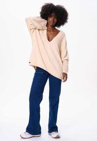 Beige oversized trui met V-hals, lange mouwen en een diagonale zoom, gecombineerd met blauwe flared jeans en witte sneakers.