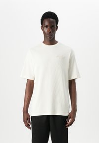 Fehér, texturált, rövid ujjú póló, kerek nyakkivágással és finom, aranyszínű logóhímzéssel a bal felső mellrészen.